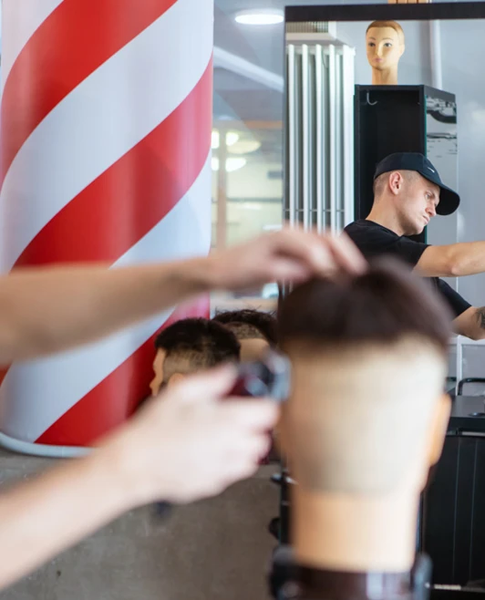 A barbering student using a spray bottle on a mannequin head while another student trims a different mannequin in the background. A barbering student using a spray bottle on a mannequin head while another student trims a different mannequin in the background.